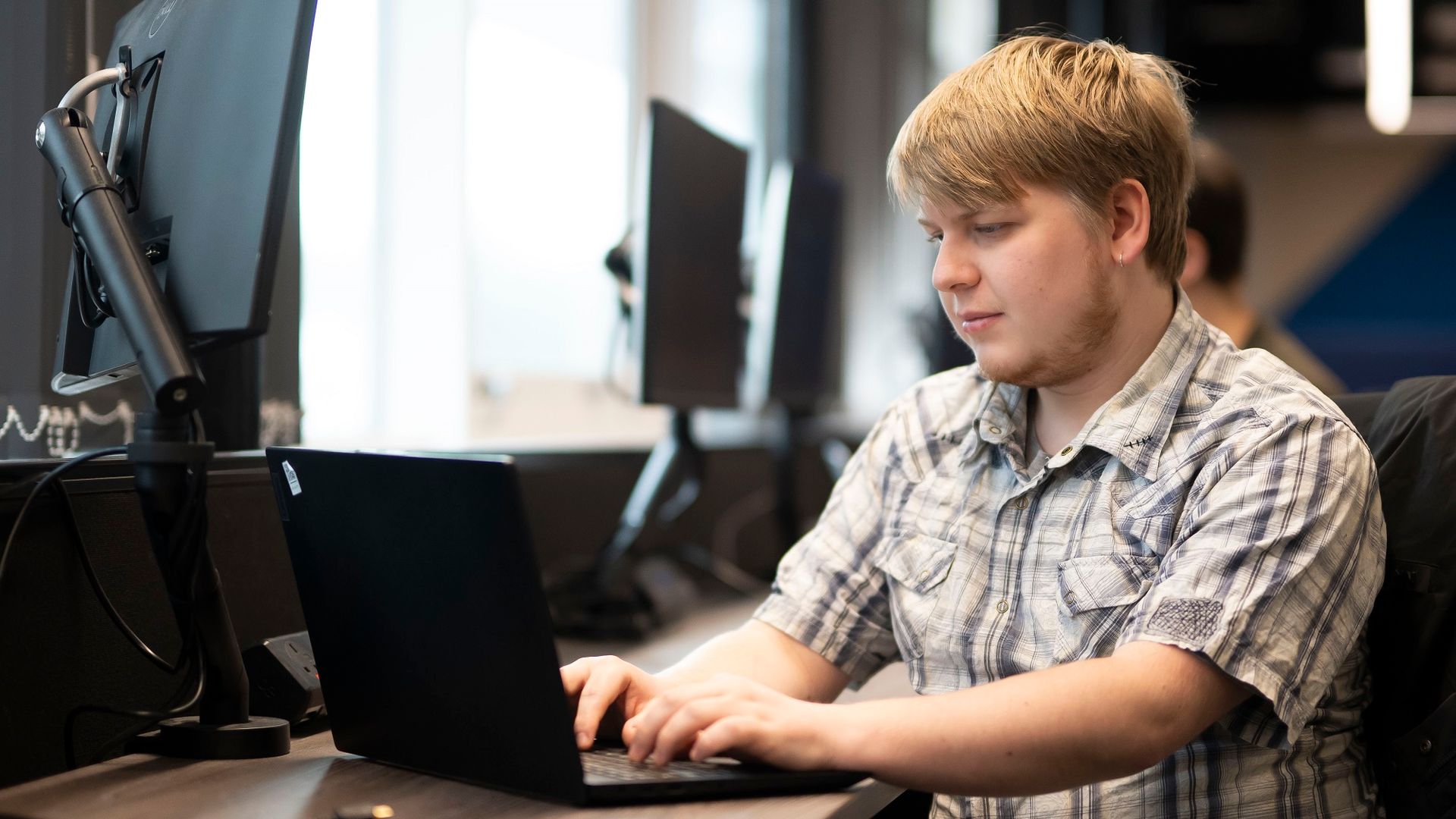 A young man in a short sleeve pattern shirt types on a laptop at a desk