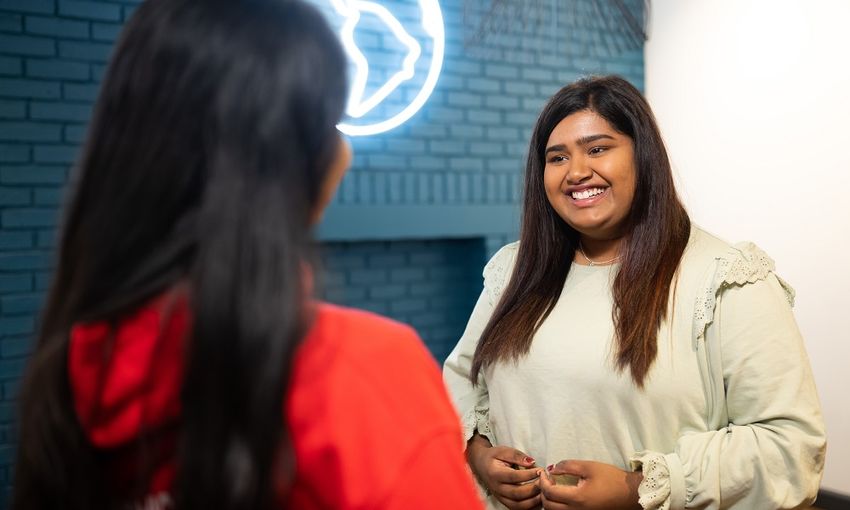A woman smiles warmly at another woman in a professional office setting, conveying a sense of camaraderie and positivity.