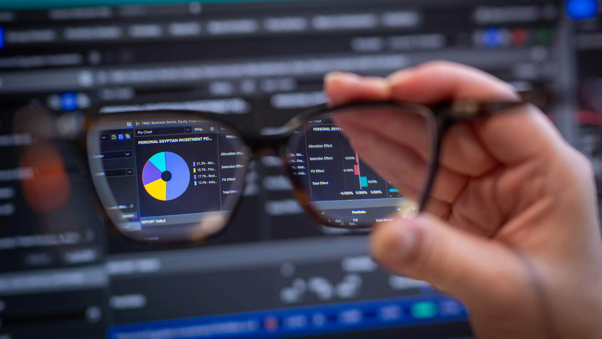 A woman with blonde hair and glasses is seated in front of multiple computer monitors displaying financial data. She appears focused and is touching her nose while working in a modern office setting.