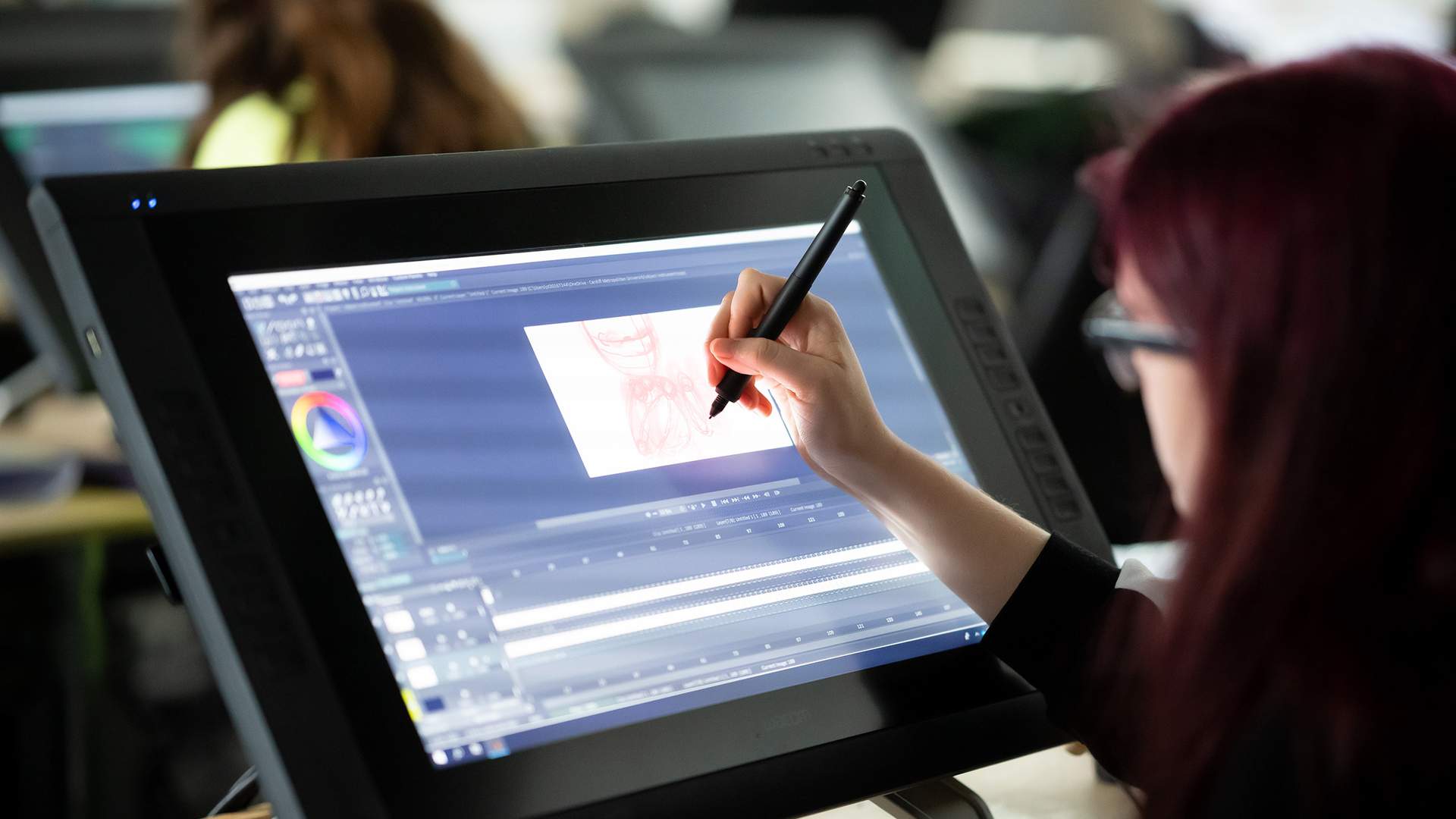 A person with long hair is using a stylus to draw on a large digital tablet. The screen displays graphic design software with artwork in progress. Other people are visible in the background, working at similar stations.