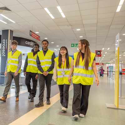 A group of five people wearing yellow safety vests walk through a corridor in an airport. They appear to be conducting an inspection or tour. The corridor is well-lit and has various signs and a glass entrance visible in the background.