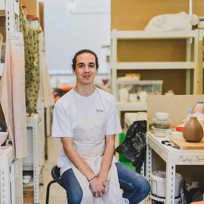 Person with long hair wearing a white apron sits on a stool in an art studio. Shelves filled with pottery, tools, and materials surround them. The environment appears creative and well-used.