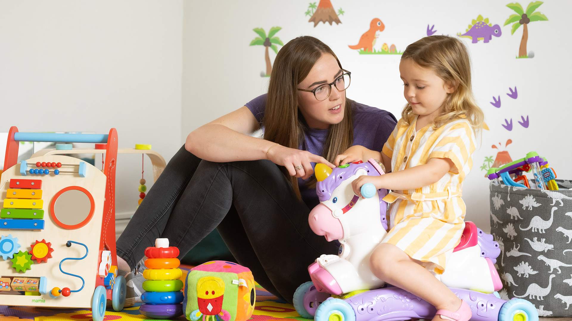 A woman with glasses plays with a young girl on a unicorn ride-on toy. They are surrounded by colorful toys and decorations, including a xylophone cart and building blocks, in a room with playful wall stickers.