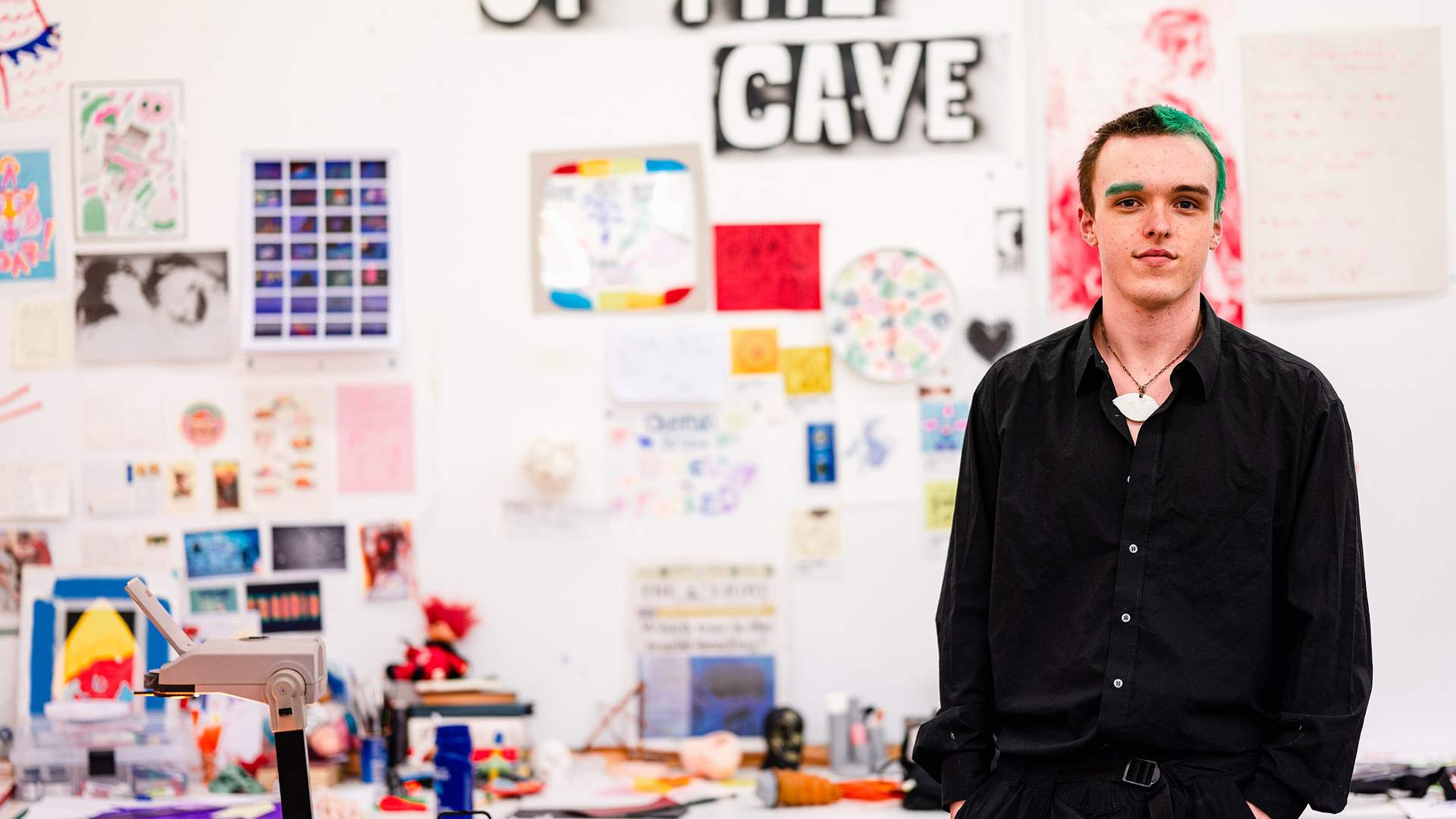 A person in a black shirt stands in a creative studio space. The wall behind them features a large OF THE CAVE sign and is covered with colorful artwork and posters. The overall atmosphere is artistic and vibrant.