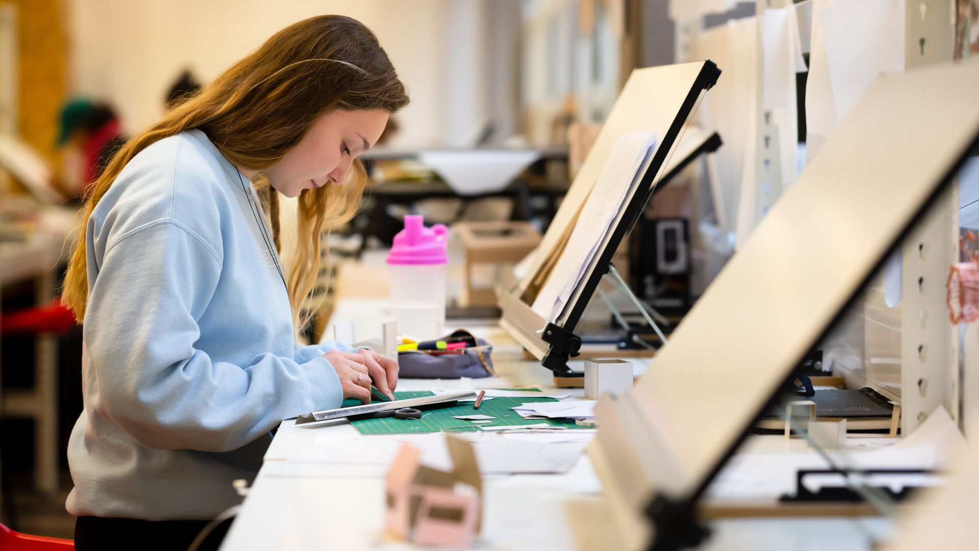 A person wearing a black shirt and black pants is focused on woodworking, marking a piece of wood with a pencil. Safety glasses rest on the table. In the background, a window and machinery are visible in a bright workshop.