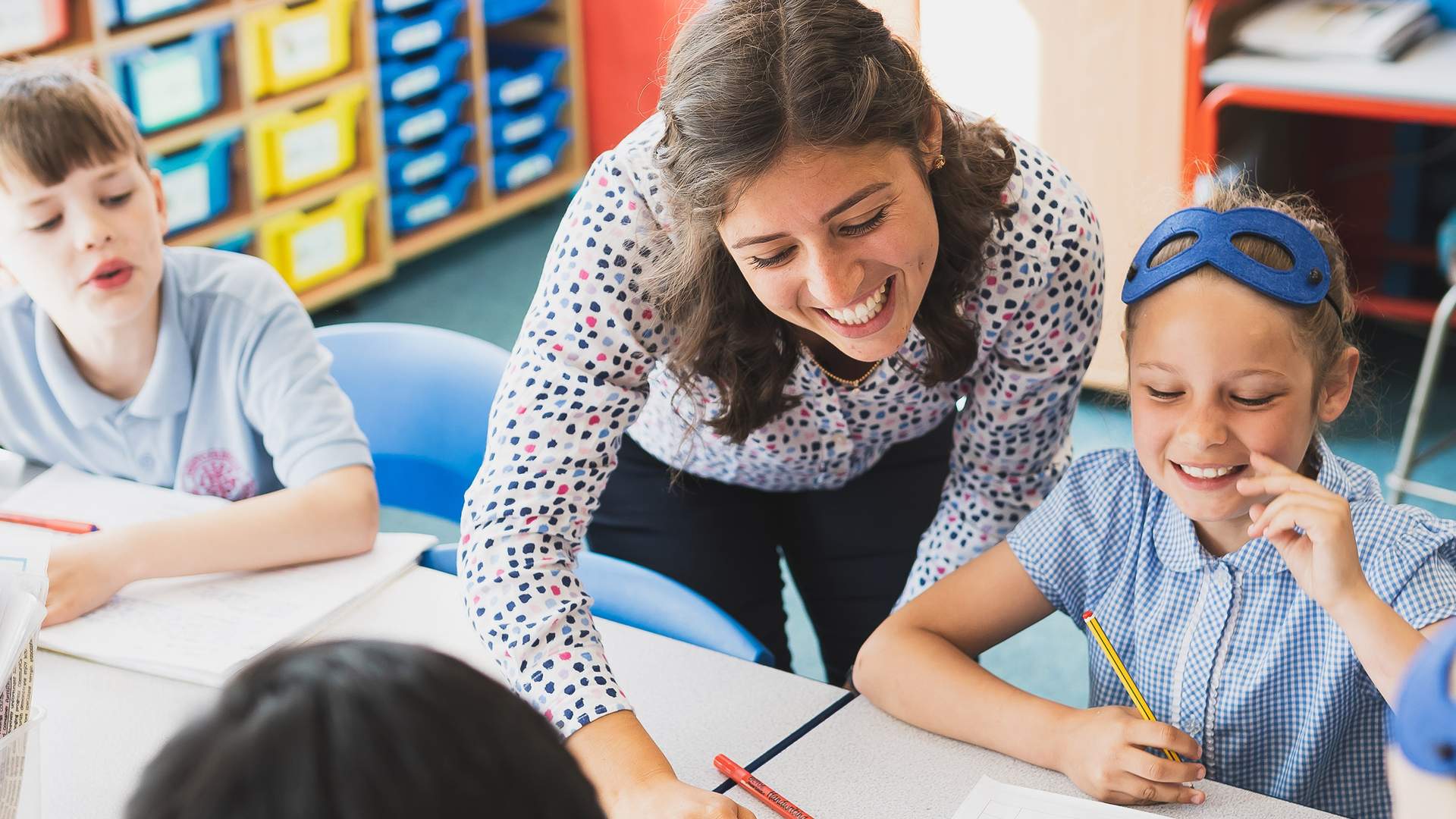 A teacher leans over a classroom table, smiling and engaging with two elementary-aged students in school uniforms. The students are seated and holding pencils, with colorful storage bins visible in the background.