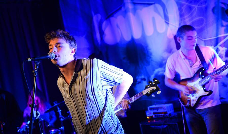 A person singing into a microphone. Behind them are a drummer and someone playing an electric guitar.