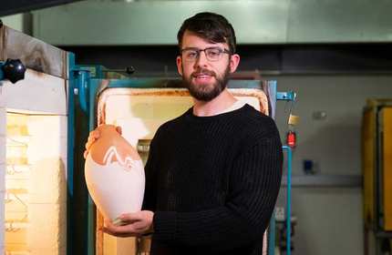 A person with a beard and glasses is holding a large ceramic vase with a white and reddish-brown pattern, standing in front of a kiln in a workshop setting. They are wearing a black sweater.