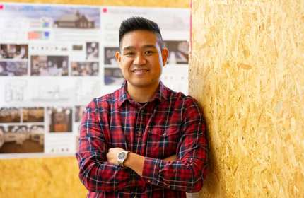 A person wearing a red plaid shirt stands with arms crossed, smiling, in front of a wooden wall. In the background, blurred posters display various images and text.