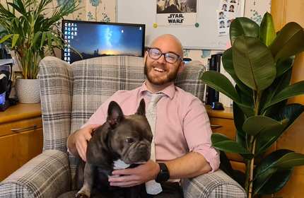 A man sits on a plaid armchair, smiling, with a French bulldog on his lap. He wears glasses, a pink shirt, and a tie. A computer, plants, and Star Wars posters are visible in the background.