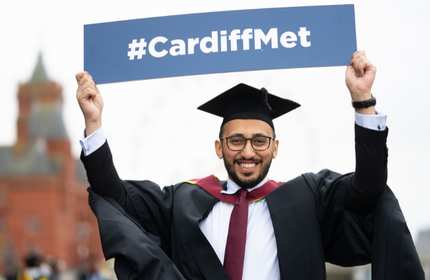Ali Salem, wearing a black graduation cap and gown, holds a #CardiffMet sign above his head