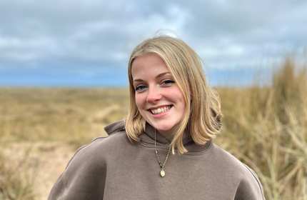 A person wearing a light brown hoodie stands in front of some sand dunes.