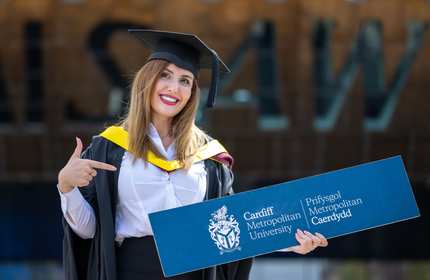 Ayda, wearing a graduation cap and gown, holds a bilingual Cardiff Met branded sign