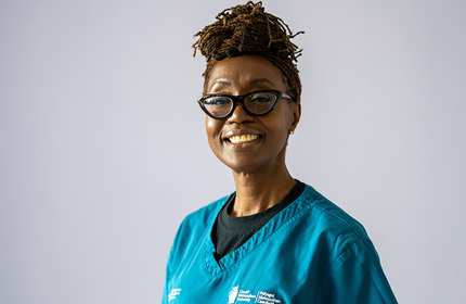 A smiling person wearing glasses and a blue medical scrub stands against a plain background. Their hair is styled in an updo.