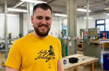 A man with a beard, wearing a yellow t-shirt, smiles in a workshop filled with woodworking tools and equipment. The background features tables, machinery, and large windows.