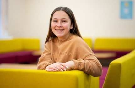 A smiling person leans on the back of a yellow chair.