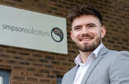 Cellan Williams, wearing a check grey suit jacket and white shirt, stands in front of a solicitors sign