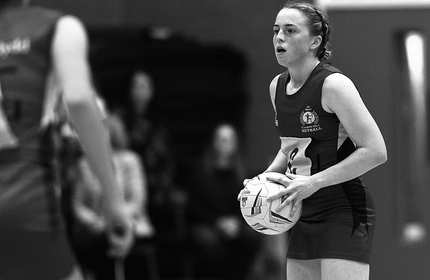 A black and white photo shows a netball player holding the ball, preparing to pass. Shes wearing a sleeveless uniform with the number 6 visible. Another player stands in the foreground. Spectators can be seen blurred in the background.