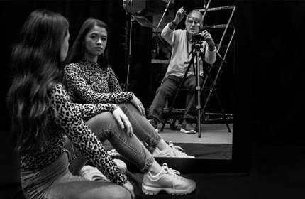A black and white photo featuring a woman sitting with her legs crossed, looking at her reflection in a large mirror. She wears a leopard print top and jeans. A photographer is visible in the mirror, taking her picture.