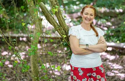 A woman with red hair stands smiling with arms crossed, leaning against a tree in a forest. She wears a white top with lace detailing and a red floral skirt. Sunlight filters through the leaves, creating dappled light on the ground.