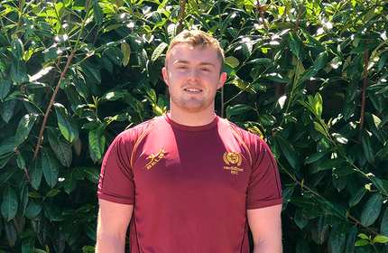 A person wearing a maroon sports jersey smiles while standing in front of a lush green hedge.