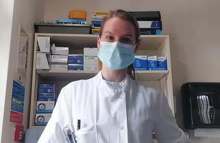 A healthcare professional wearing a white coat and a blue surgical mask stands in front of shelves stocked with medical supplies and gloves. The setting appears to be a clinic or a hospital.