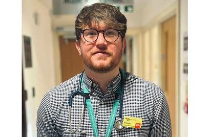 A person with glasses and a beard stands in a hallway wearing a checkered shirt, a green lanyard, and a stethoscope around their neck. They have a name badge clipped to their shirt.
