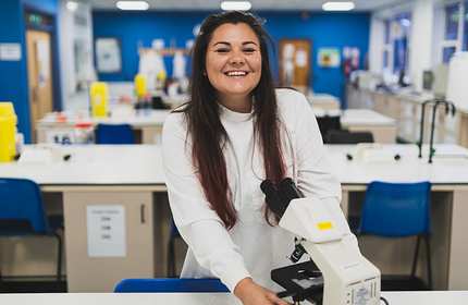 A woman wearing a lab coat smiles while standing in a science laboratory. She is leaning on a microscope with tables and lab equipment visible in the background. The room has blue walls and bright lighting.