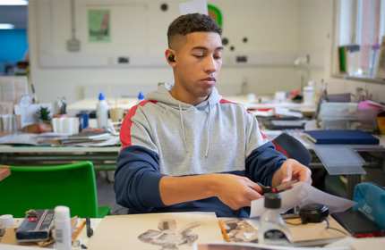 A person sits at a desk, covered with drawings, books and pens. They are cutting a piece of paper with a scissors.