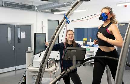 A person runs on a treadmill while wearing a breathing mask. A second person stands alongside, next to a computer display and other equipment.