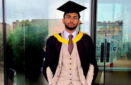 A graduate stands in front of a glass building, wearing a traditional cap and gown with a yellow and black hood. The graduate also has a beige plaid suit and a brown tie. The background features trees and urban elements reflected in the glass.