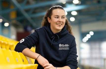 A woman with long hair is sitting in a yellow stadium seat, wearing a navy hoodie. She is smiling and looking to the side. The background shows a blurred indoor sports facility.