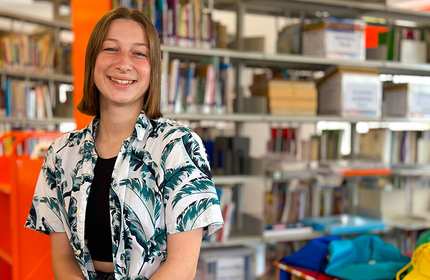 A smiling person stands in a library wearing a tropical-patterned shirt over a black top. Shelves filled with books and colorful bins are visible in the background, creating a vibrant and welcoming atmosphere.