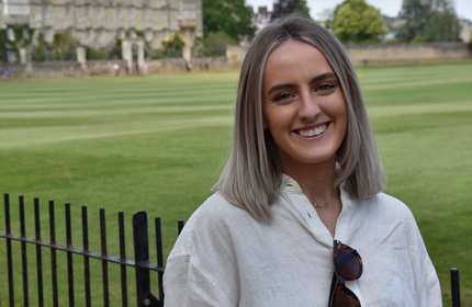 A person stands in front of railings. In the background is a large area of grass.