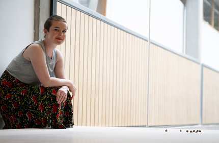 A person sits against a wall. Their arms are folded on a raised knee.