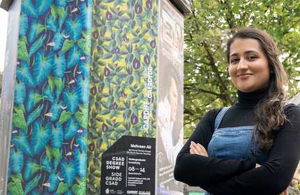 A woman stands confidently with crossed arms in front of a vibrant art installation featuring colorful leaf patterns. Shes wearing a black turtleneck and denim overalls. A tree and part of a building are visible in the background.