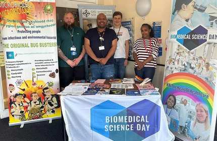A group of five people stand behind a table covered with brochures and posters. The tablecloth reads Biomedical Science Day. Banners about microbiology and healthcare flank the group. They smile in a brightly lit room.
