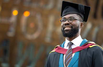 Ismaila Ibrahim, wearing a black graduate cap and gown, stands in front of the Cardiff Millennium Centre