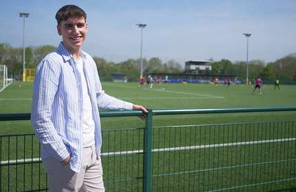 A person stands in front of low metal railings. In the background is a playing field.