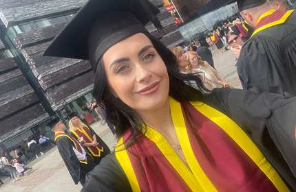 Kirsty Bennetta, wearing a black graduation cap and gown and a red and yellow sash, takes a selfie in front of other groups of graduates during Graduation day