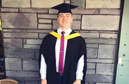 A man wearing academic robes over a shirt and tie stands in front of a wall.