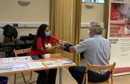 A healthcare worker in a red shirt checks a mans blood pressure at a clinic. Both are wearing masks. A poster promoting regular health checks is visible in the background. A table with pamphlets and health supplies is nearby.