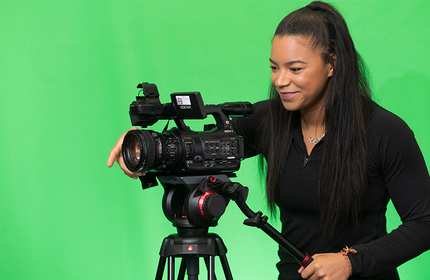 Mica Moore operates a video camera on a tripod in front of a large green screen
