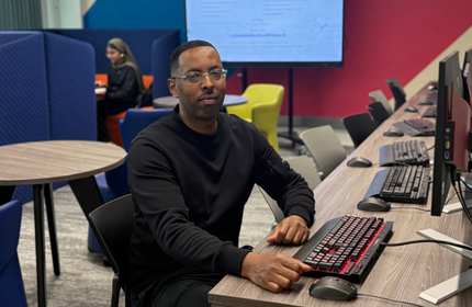 A person sits at a computer workstation. In the background are several other workstations and a colourful seating area.