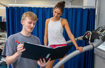 A woman in athletic wear stands on a treadmill, looking at a man holding a black folder. The man, in a gray shirt, is writing notes. They are in a gym setting with blue curtains and fitness equipment visible in the background.