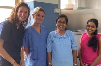 Four women stand together in a room with a window. Two are wearing blue medical scrubs, and two are in casual clothes. They are smiling at the camera. The setting appears to be a medical or healthcare facility.