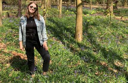 A woman stands in a sunlit forest, wearing sunglasses, a denim jacket, and black pants. She smiles while surrounded by tall green plants and scattered blue flowers. Sunlight filters through the trees, casting dappled shadows.