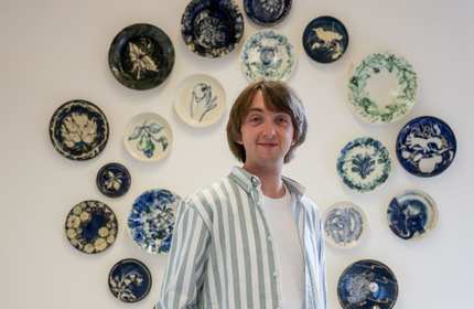 A man stands confidently in front of a wall adorned with various decorative plates, showcasing a unique collection.
