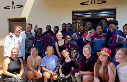 A diverse group of people, including men and women, pose together outside a building. Some hold red and green flags. Many are smiling and casually dressed, with a mix of athletic and casual outfits. The building has a beige exterior.