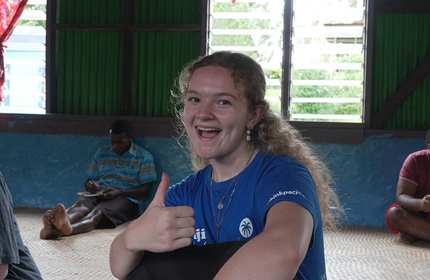 A person sitting on a matted floor gives a thumbs up, smiling at the camera.